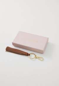 Porte-clés en cuir marron avec un pompon et des accessoires dorés. Accompagné d'une boîte rose clair avec un texte doré.