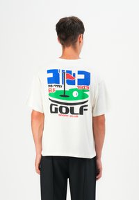 Lyhythihainen, valkoinen puuvillapaita, jossa on värikäs graafinen painatus golfskenesta. Tekstissä lukee "GOLF," "SPORT CLUB," ja japaninkielisiä merkkejä.