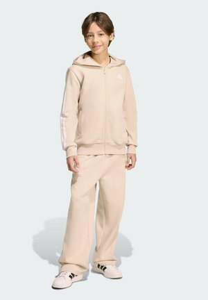 Sudadera beige con capucha, cierre frontal y franjas blancas en las mangas, combinada con pantalones holgados a juego y zapatillas blancas.