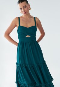 Abito turchese con spalline sottili, corpetto intrecciato con una apertura e gonna a balze con dettagli arricciati. Tessuto dalla texture liscia.