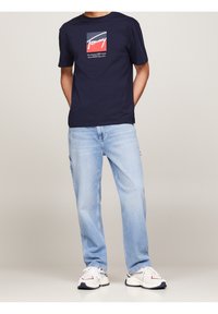 Námořnicky modré tričko s krátkým rukávem a logem, doplněné světlými modrými džíny a bílými teniskami s červenými a námořnickými detaily.