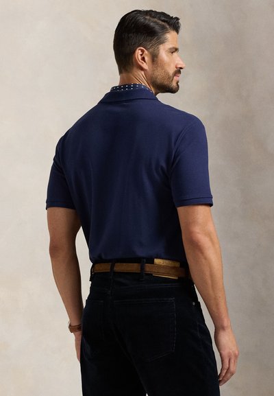Μπλε ναυτικό πόλο πουκάμισο με μοτίβο στο γιακά, κοντά μανίκια και ά νετη εφαρμογή, συνδυασμένο με σκούρο τζιν και ταμπά ζώνη. Λείο υφασμάτινο υλικό.