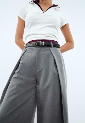 Persona che indossa dei pantaloni grigi a vita alta e ampia con una cintura nera e una maglietta bianca a maniche corte a costine, con le mani dietro la schiena.