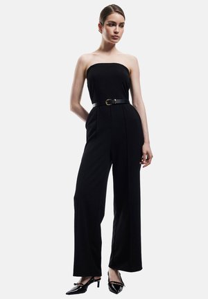 Zwarte strapless jumpsuit met een aansluitend lijfje en wijde broekspijpen. Heeft een zwarte riem met een gouden gesp en glanzende zwarte hakken.