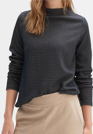 Femme portant un haut texturé à manches longues foncé, rentré dans un pantalon beige taille haute, debout devant un fond uni.