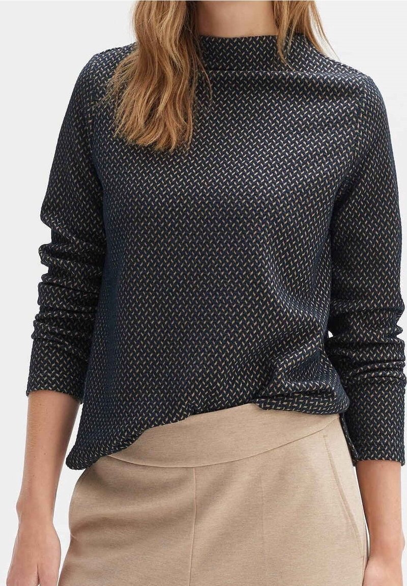 Femme portant un haut texturé à manches longues foncé, rentré dans un pantalon beige taille haute, debout devant un fond uni.