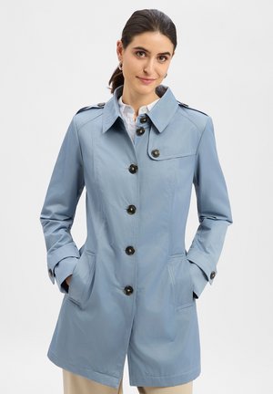 Frau trägt einen hellblauen, geknöpften Trenchcoat mit den Händen in den Taschen, darunter ein weißes Hemd, und die Haare sind zurückgebunden vor einem schlichten Hintergrund.