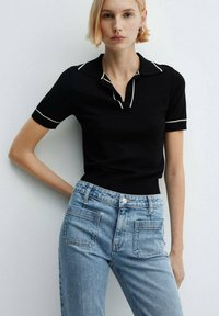 Una joven con cabello corto rubio lleva una camiseta negra de manga corta con ribete blanco y jeans de tiro alto de color azul claro, posando frente a una pared blanca.