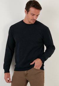 Sötétkék, crewneck pulóver, puha anyagból készült, bordázott mandzsettákkal és alsó szegéllyel, laza illeszkedéssel. Barna nadrággal látható.