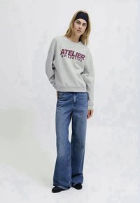Sweatshirt cinza com o texto "ATELIER COLLECTIVE" em bordô, combinado com jeans de perna larga azuis e sapatos pretos, em pé num fundo neutro.
