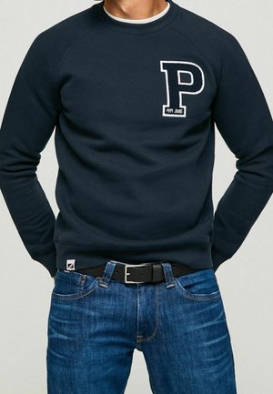 Mann trägt einen marineblauen Pullover mit großem „P“ und dem Schriftzug „Pepe Jeans“ auf der Brust, kombiniert mit blauen Jeans und schwarzem Gürtel.