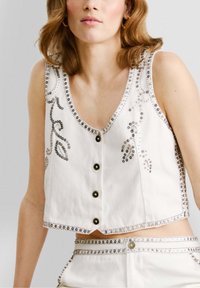 Bijela skraćena majica bez rukava s dekorativnim srebrnim nitnama, dubokim V-izrezom i crnim kopčama. Detaljno ukrašavanje perlama duž rubova.