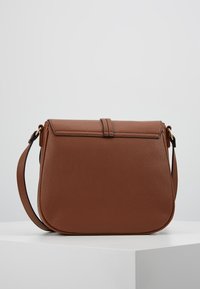 Ruskea nahkainen olkalaukku, jossa on läppäkiinnitys, pyöristetyt reunat ja säädettävä olkahihna, sijoitettuna valkoiselle pinnalle neutraalia taustaa vasten.