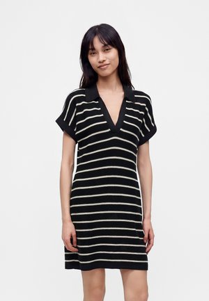 Joven mujer con cabello lacio y oscuro lleva un vestido negro de manga corta con rayas horizontales blancas y un escote en V profundo.