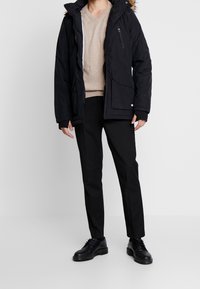 Henkilö, joka on pukeutunut mustaan talvitakkiin, jossa on turkisvuorattu hupunsä ja beige pusero, mustiin housuihin ja mustiin nahkasandaaleihin, seisoo yksinkertaisen valkoisen taustan edessä.