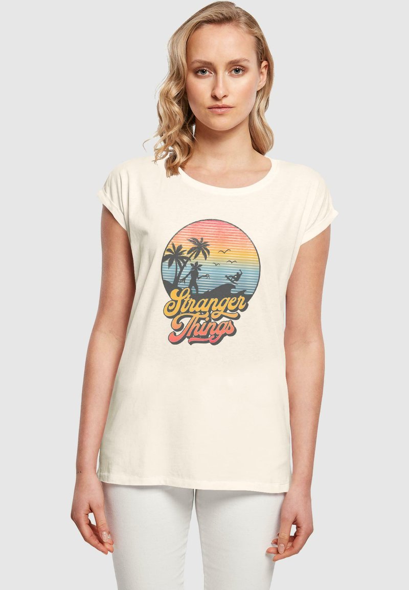 Cremefarvet bomulds t-shirt med et cirkulært grafik, der har palmetræer, en solnedgangsgradient og teksten "Stranger Things" i livlige farver.
