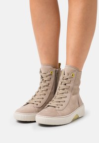 Baskets montantes en suède beige clair avec un système de laçage à l'avant, œillets jaunes, fermeture éclaire latérale et semelle en caoutchouc blanc texturé avec un accent jaune.