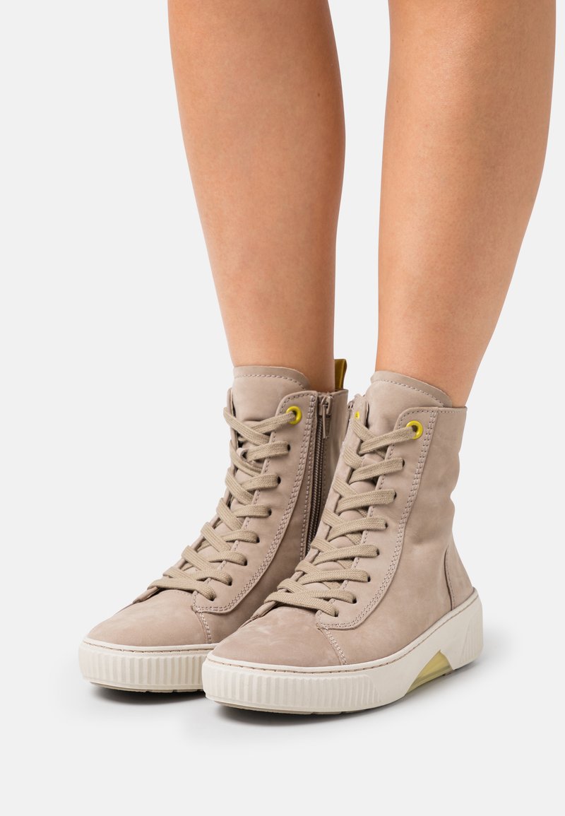 Baskets montantes en suède beige clair avec un système de laçage à l'avant, œillets jaunes, fermeture éclaire latérale et semelle en caoutchouc blanc texturé avec un accent jaune.