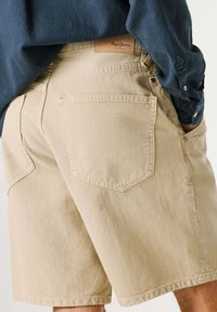 Person wearing beige denim shorts with back pockets and a dark blue long-sleeve shirt, right hand in pocket, brand label visible.