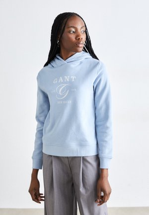 Ανοιχτό γαλάζια φούτερ με κουκούλα και μπροστινό λογότυπο, ριχτές μανσέτες και στρίφωμα. Φοριέται με ανοιχτό γκρι πλισέ παντελόνι.