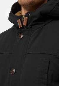 Veste noire à capuche avec un intérieur texturé, comprenant des accents en cuir marron, de gros boutons et une fermeture éclair solide.