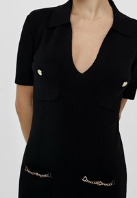 Svart kortärmad V-ringad klänning med krage, två bröstfickor med guldknappar och två nederfickor med guldkedjedetaljer.