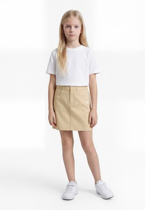 Noor tüdruk seisab ettepoole vaadates, kandes valget t-särki, beeži logo ja valgete tossudega ühetasandilisel taustal.