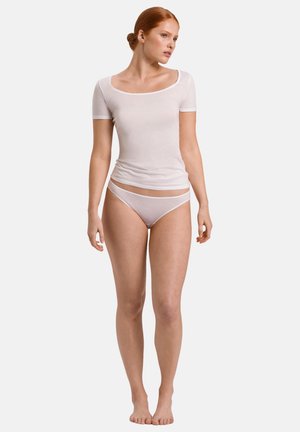 Nuori nainen punaisilla hiuksilla, yllään istuva valkoinen lyhythihainen paita ja valkoiset alusvaatteet, seisoo paljain jaloin yksiväristä taustaa vasten.