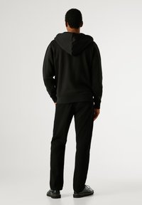 Μαύρη βαμβακερή φούτερ με μεγάλη κουκούλα, ραμμένα μανίκια και ύφασμα στο τελείωμα. Συνδυάζεται με μαύρα παντελόνια που ταιριάζουν. Ο μοντέλο στέκεται με τα χέρια στις τσέπες.