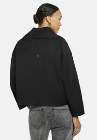 Vrouw met haar in een knot die gouden oorringen draagt, een zwarte cropped jacket en grijze gewassen jeans, van achteren gezien en naar rechts kijkend.