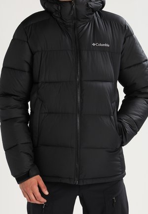 Sort Columbia isoleret dunjakke med lynlås foran og hætte, båret med hænderne i lommerne og ensfarvet lys baggrund.