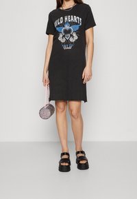 Musta T-paita mekko, jossa on graafinen printti "Wild Hearts Club", yhdistettynä paksupohjaisiin mustiin sandaaleihin ja pieneen, pyöreään, kimaltelevaan käsilaukkuun.