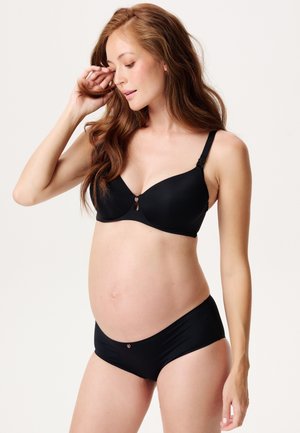 Gravid kvinde med langt hår iført sort amme-bh og matchende undertøj, stående mod en simpel lys baggrund.