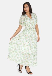 Robe fleurie vert clair avec des manches courtes à volants, une taille cintrée et une longueur maxi fluide, associée à des talons avec brides à la cheville de couleur noire.