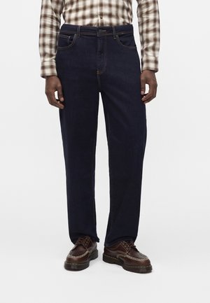 Homme portant un jean bleu foncé à jambes droites, des chaussures en cuir marron à lacets avec coutures blanches, et une chemise à carreaux beige à manches longues.