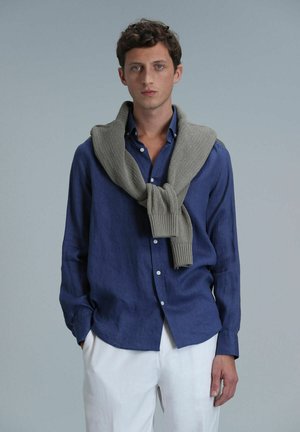 Chemise en lin bleu marine à manches longues, boutonnée, associée à un pull en tricot gris clair drapé sur les épaules, et un pantalon blanc.