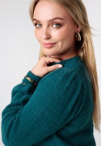 Groenblauw, pluizige trui met een ronde halslijn en een zachte textuur; gecombineerd met gouden armbanden en oorbellen. Model lacht en kijkt in de camera.