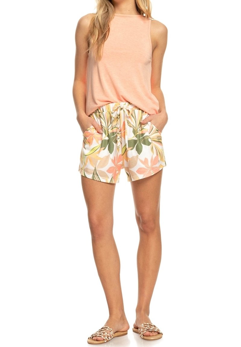 Femme portant un haut sans manches pêche, un short à cordon fleuri et des sandales tressées à bouts ouverts, debout avec les mains dans les poches.