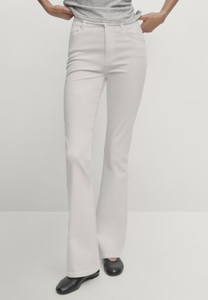 Personne portant un pantalon évasé blanc, une chemise grise et des chaussures noires, debout les mains près des poches sur un fond uni.