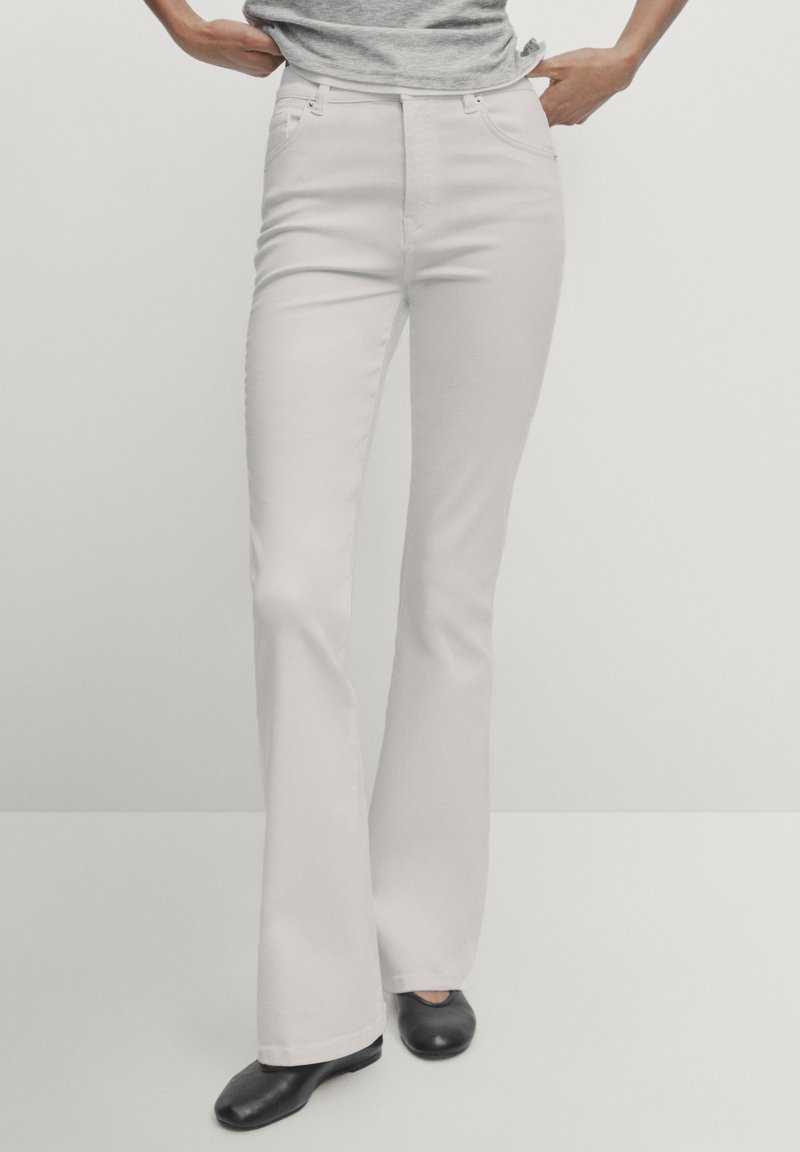 Personne portant un pantalon évasé blanc, une chemise grise et des chaussures noires, debout les mains près des poches sur un fond uni.