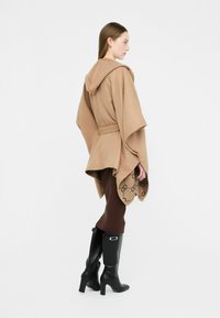 Poncho marrone con cappuccio e cintura, caratterizzato da una texture liscia e un bordo decorato. Abbinato a leggings marroni e stivali neri con tacco al ginocchio.