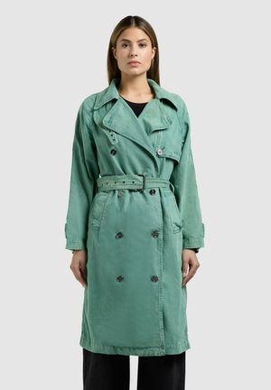 Frau steht mit dem Gesicht nach vorne und trägt einen zweireihigen, taillierten Trenchcoat in Türkis über einer schwarzen Hose vor einem schlichten Hintergrund.