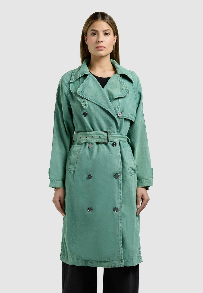 Nő áll szemben, aki zöldeskék, dupla gombos, övvel ellátott ballonkabátot visel fekete nadrág fölött, egyszerű háttér előtt.