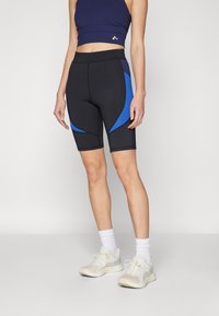 Pantaloni neri con pannelli laterali blu, design aderente e texture liscia; abbinati a un crop top blu navy e scarpe da ginnastica chiare.