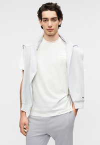 Άσπρο μπλουζάκι με υφασμένη ετικέτα λογότυπου, σε συνδυασμό με ένα ανοιχτό μπλε φούτερ με φερμουάρ, κρεμασμένο στους ώμους. Γκρι παντελόνι με λεπτομέρειες.