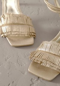 Gouden sandalen met een vierkante neus, gelaagde franjes en een gladde metalen afwerking op een gestructureerd oppervlak.