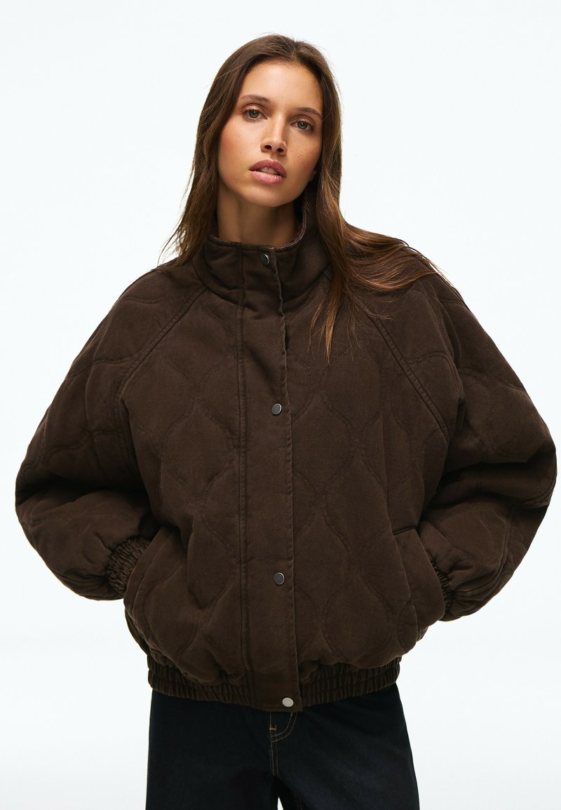 Jeune femme portant une veste matelassée marron foncé avec des boutons-pression, debout les mains dans les poches devant un fond uni.