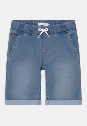 Jasnoniebieskie szorty jeansowe z elastycznym pasem, białym ściągaczem, podwiniętymi nogawkami i bocznymi kieszeniami, ułożone płasko na białym tle.