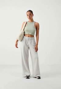 Kvinna som står och bär en ljusgrön croppad tanktop, ljusgrå vida sweatpants, svartvita sneakers och har en beige axelväska.