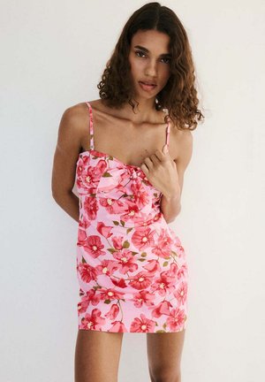 Mujer con cabello rizado que lleva un vestido sin mangas rosa con flores y tirantes finos, posando frente a un fondo blanco liso.